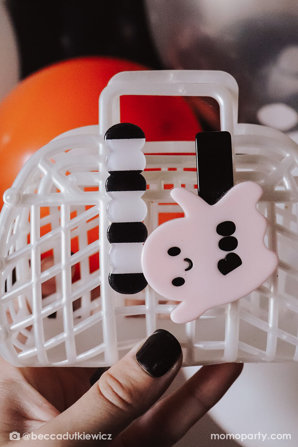 a hand holding a mini white jelly basket with a super cute Ghost Light Pink + Wave Black Stripe Alligator Hair Clips on it, in front of Meri Meri Halloween Balloon Garland, mixed with black, silver, orange, white and confetti balloons and hanging bat decoration. simple and cute Spooky Basket Idea That Are All Treat, No Tricks for your little Boo