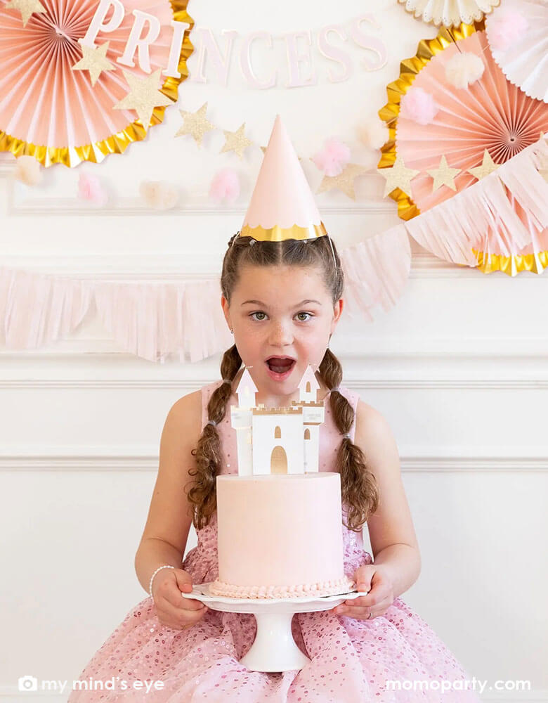 A Girl holding a princess themed birthday cake standing in front of a wall decorated-with-princess-themed-decorations