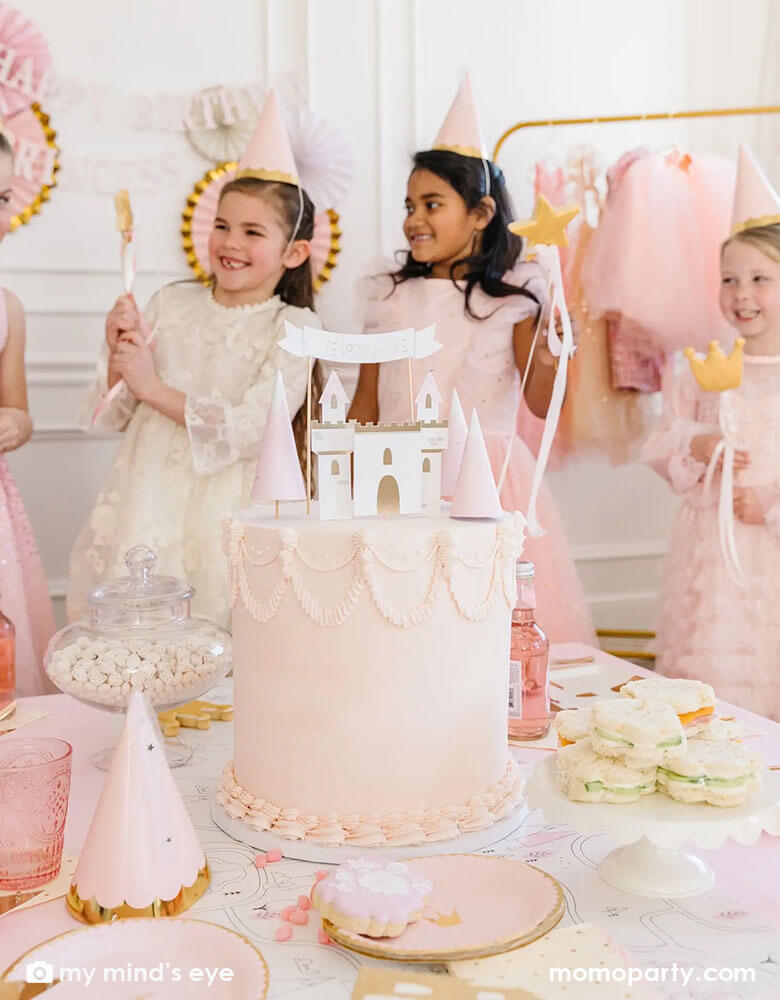 A group of girls dressed up as princesses in princess party hats and star wands standing in the back of a gorgeous princess themed pink birthday cake decorated with My Mind's eye cake topper set featuring pieces of castles, mountains, and "once upon a time" banner
