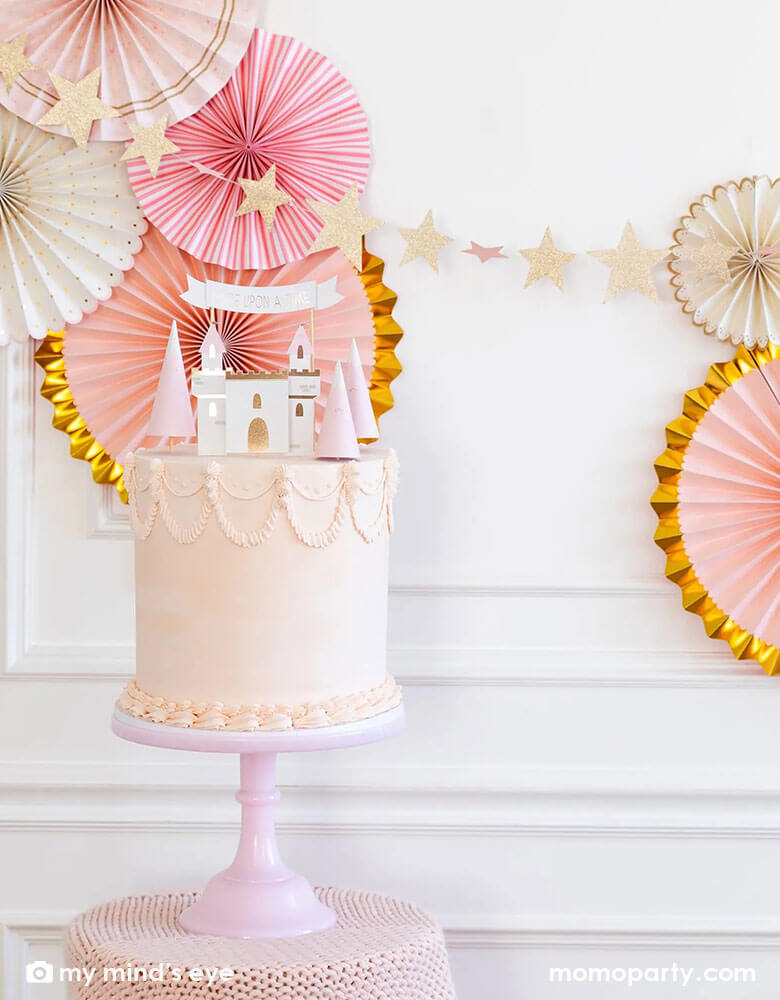 A gorgeous princess themed birthday cake decorated with My Mind's eye Cake topper set featuring castle pieces, mountains and a "Once Upon a Time" banner that will make any cake a the perfect addition to any princess party. Behind the cake is a beautiful wall decorated with princess themed party fans and banner in pretty pink and gold, a perfect inspo for a girl's princess themed birthday party