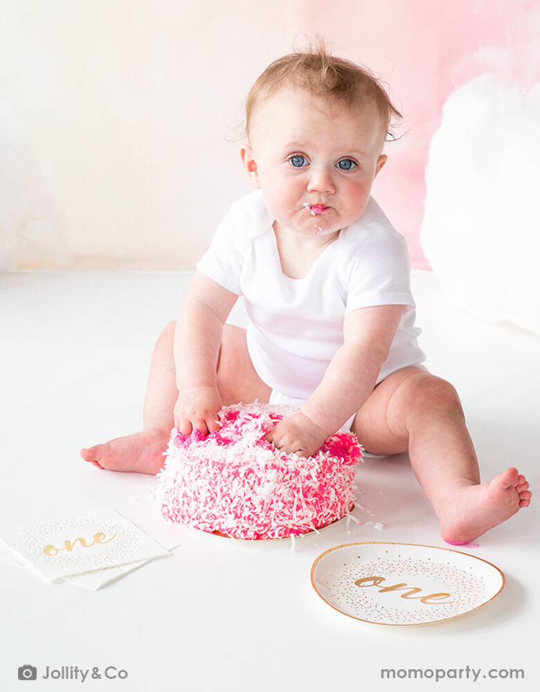 A baby's smash cake first birthday celebration with  Jollity 7-inch onederland gold round dessert plate with gold script "one" on it and gold foil confetti illustration around it with coordinated Jollity 5 inch onederland small napkins spread around 