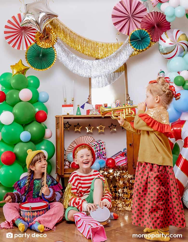 Kids dressed up in their festive outfits celebrating Christmas with a balloon Christmas tree in the back, paper fans in the classic Holiday colors of green and red, along with silver and gold tinsel garlands in hung on the wall, and candy cane foil balloons on the side, all makes great Christmas decorations this Holiday season.