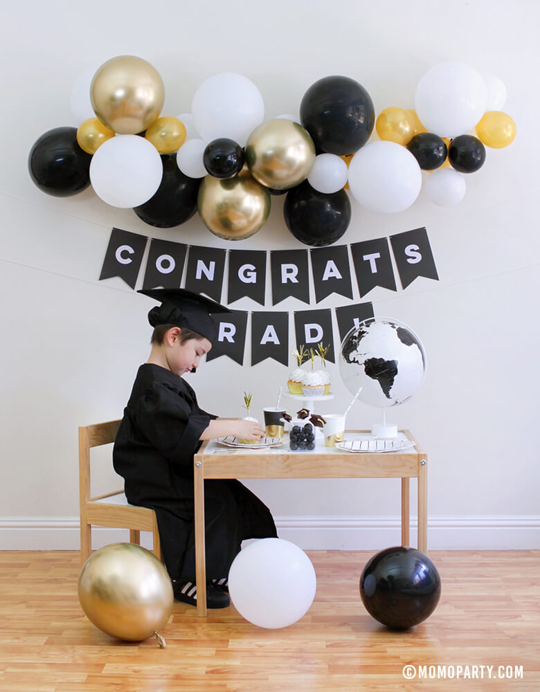 2020 Graduation at home celebration with boy siting in from the a desert table with cupcakes, black stripe plates, graduation hat lollipop,  black and white globe, books and Black banner with "Congrats Grad!" sign and Balloon garland with gold, black, white as wall decoration