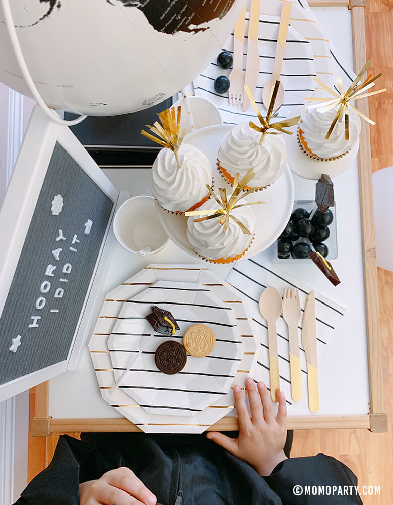modern graduation party at home idea with boy wear a black graduation gown siting in front of his table with black and gold stripe plates, napkins, Assorted Gold Dipped Cups, cupcake, cookies, b&w globe, letter board with "hooray i did it" sign, black gum balls