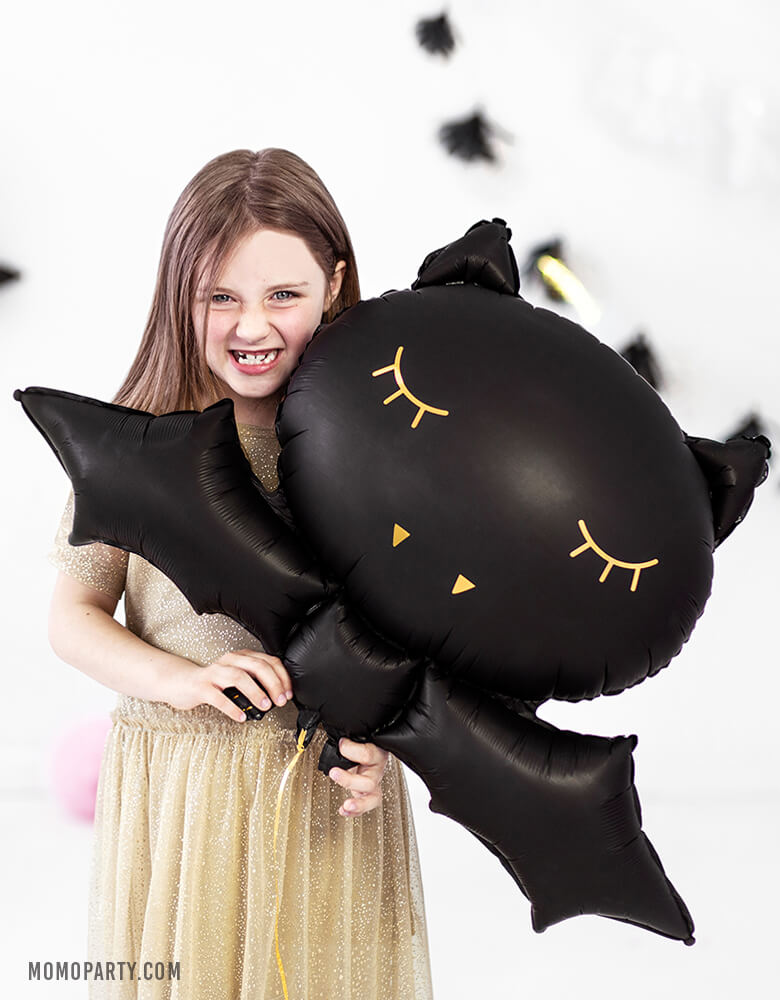 Girl holding a Party Deco 32 inches Halloween Black Bat Foil Mylar Balloon, with gold print on the black bat shape. for a kids modern spooky halloween birthday party. this balloon also perfect for hocus pocus party trick-or-treating party, nightmare before christmas party, witch themed party and all halloween related celebrations