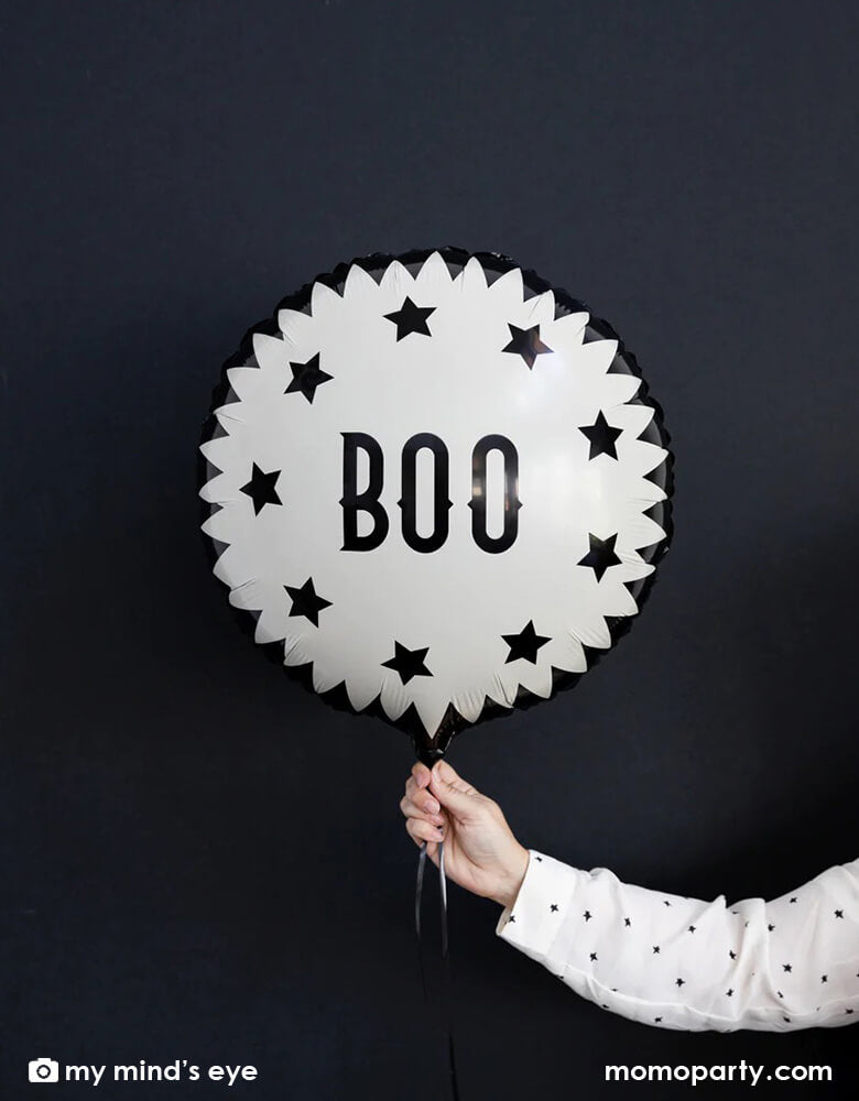 A woman's hand with white polka dot shirt holding My Mind's Eye Vintage Halloween Boo Mylar Balloon against black background creating a spooky vibe for a Halloween celebration