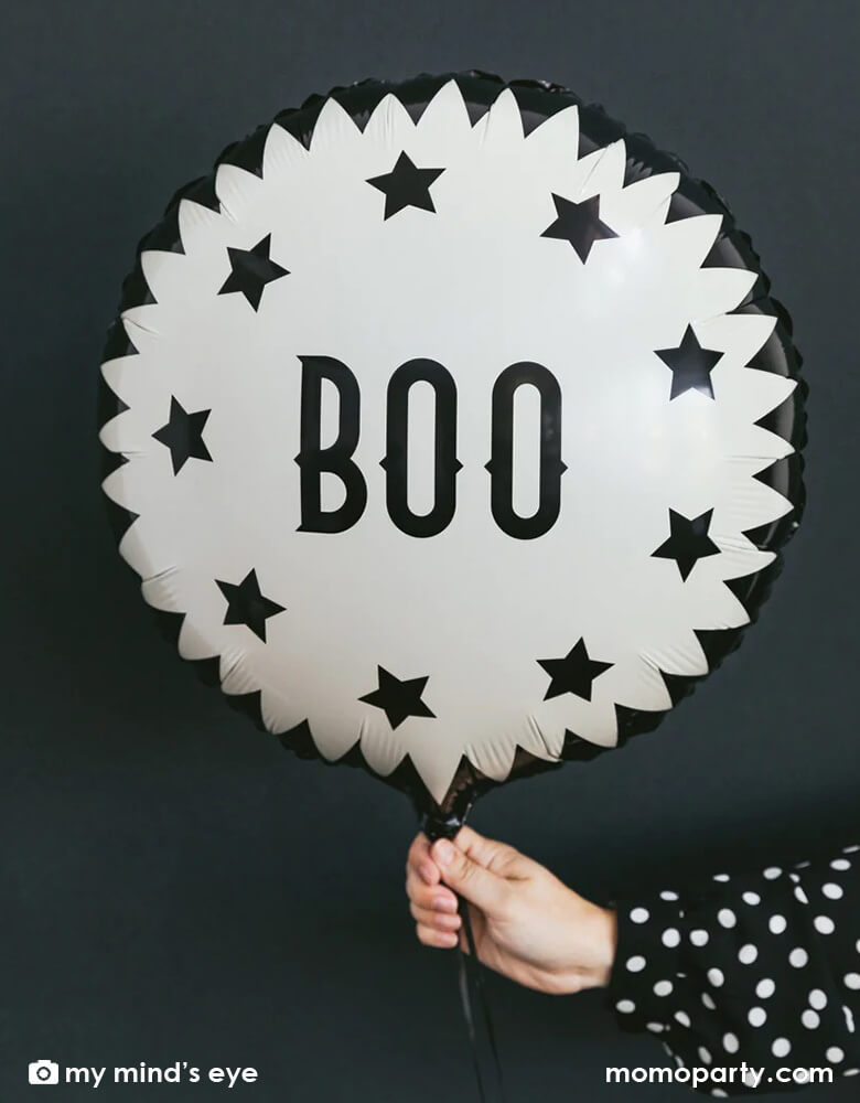 A woman's hand with black polka dot shirt holding My Mind's Eye Vintage Halloween Boo Mylar Balloon against black background creating a spooky vibe for a Halloween celebration