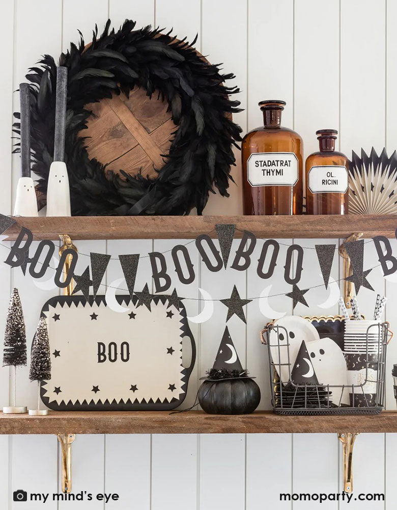 A kitchen shelf decorated with Halloween decorations including My Mind's Eye Vintage Halloween bamboo tray, witch party hats, Ghost shaped plates and napkins and Vintage Halloween Boo Banner set in black and white, with a crow feather wreath and witch potion bottles, all together creating a spooky vibe for a Halloween celebration