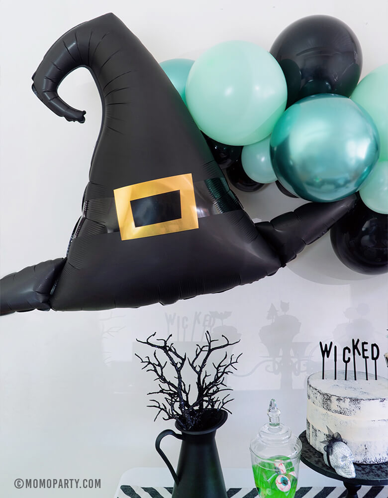 Close up look of Momo Party Halloween collection, Witch Please Party, Anagram 39" Halloween Witch Hat Satin Foil Balloon and Pastel Matt Green, Chrome Green, and Black color mixed Latex Balloon Garland as wall decoration, black pot with black branch, white cake with letterboard cake topper spelled "Wiched", Apple jelly with eyeball candy in a Jar. Creep cool look for A Modern Witch Inspired Halloween Party