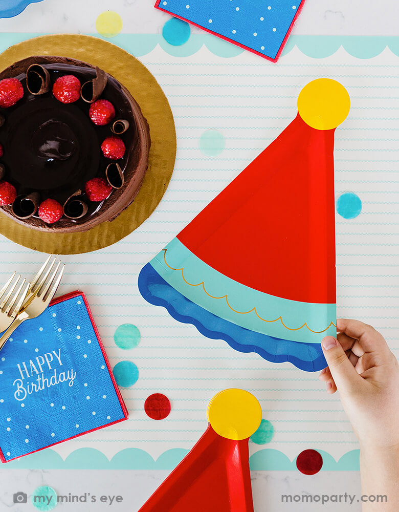 A festive birthday party table with a chocolate birthday cake, filled with cheerful confetti in blue, red and yellow and My Mind's Eye blue Happy Birthday napkins with blue birthday hat shaped party plates - a perfect size for serving birthday cake - great modern and simple ideas for a boy's birthday celebration.