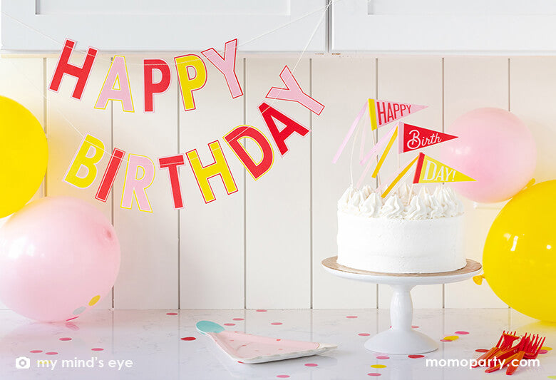 A simple and modern at-home birthday party decoration set up for a girl's birthday party at the kitchen counter space with pink and yellow birthday party balloons, My Mind's Eye's pink birthday Banner in cute colors of pink coral, yellow and red red, yellow, with a birthday cake topped with My Mind's Eye Happy birthday pink pennant cake toppers with cheerful ribbons in darling pink and peach. Perfect for a girly themed birthday party!