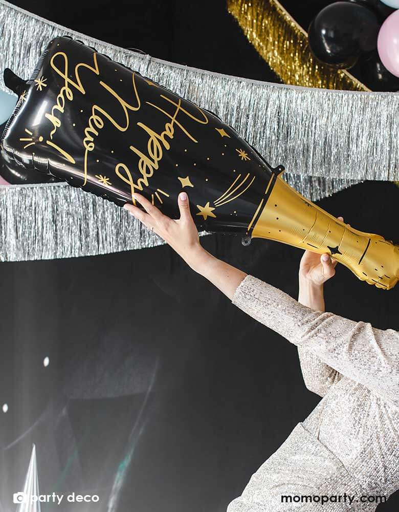 A young lady holding Momo Party's 34.5" champagne bottle shaped foil balloon in gold and black by Party Deco, standing in front of a backdrop all decorated with gold and silver tinsel garland,  a perfect decoration idea for New Years Eve countdown party!