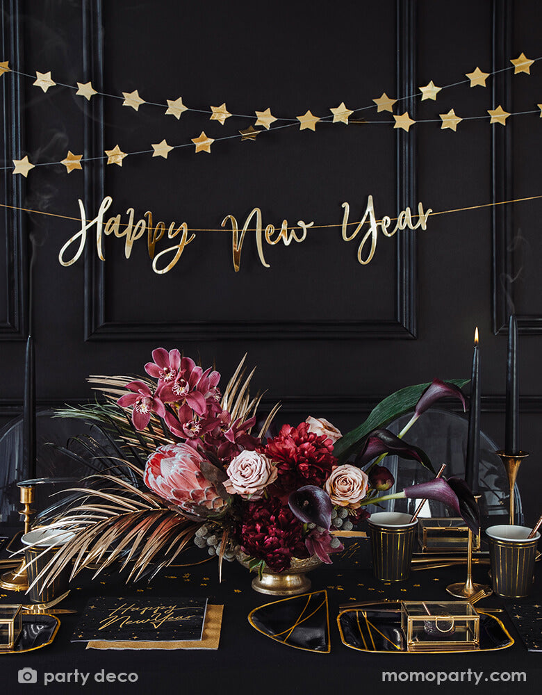 Black and gold new year party table set up with Party Deco Happy new year gold script letter banner and gold start banners hanging on the black wall, Happy New Year Large Napkins layered with gold napkin, black and gold foil special shaped party plates, cups, gold star confetti, candles and beautiful Floral arrangement in a gold vase as centerpiece in a black table top. Ring in the New Year in style with this gorgeous Happy New Year party collection with gold foil detail!