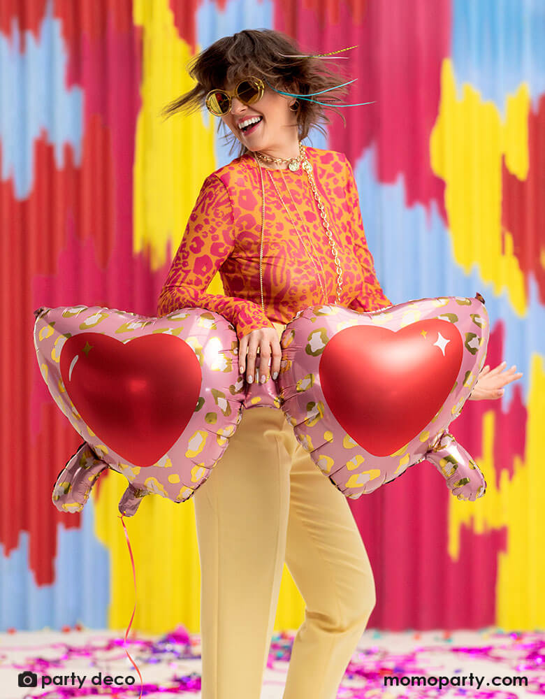 A young lady in her leopard shirt holding Momo Party's 30 inch heart shaped sunglasses foil balloon by Party Deco, With animal print design around the glasses in pink and red colors in her arms, standing in front of a colorful bright wall, celebrating love for Valentine's Day.