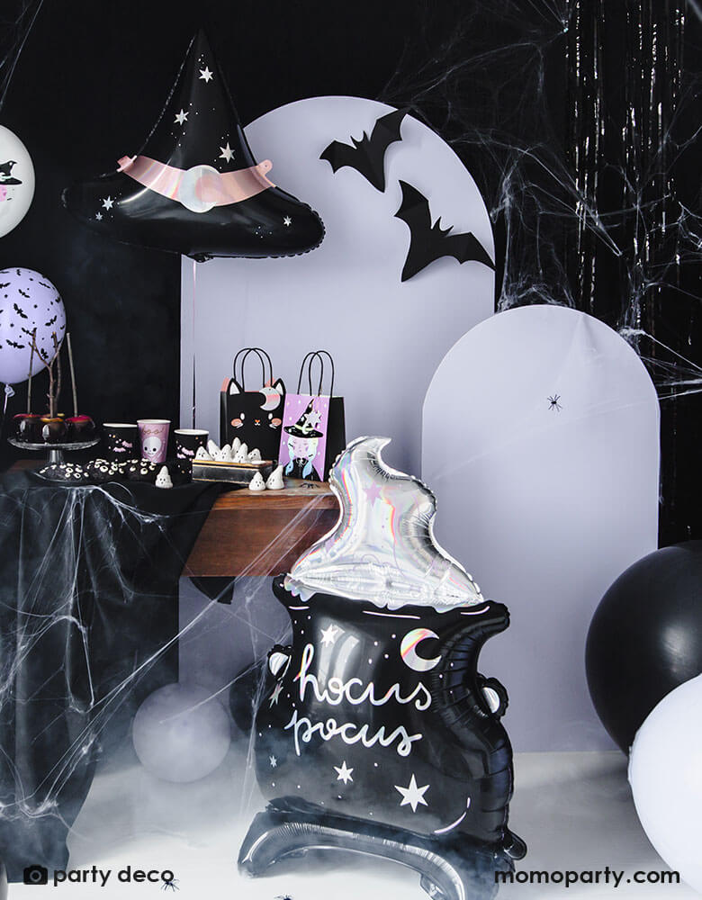 A spooky cute Hocus Pocus themed Halloween party set up featuring Momo Party's witch themed party supplies including the Hocus Pocus Cauldron shaped standing foil balloon, Boo! party cups in pink and blakc, witch party bags on the table. In the back is a lilac wooden backdrop adorn with Momo Party's 19" witch hat shaped foil balloon and bat paper decoration, with black and white balloons on the floor and spiderweb stretched to the table and backdrop, all makes fun and festive Halloween decors!