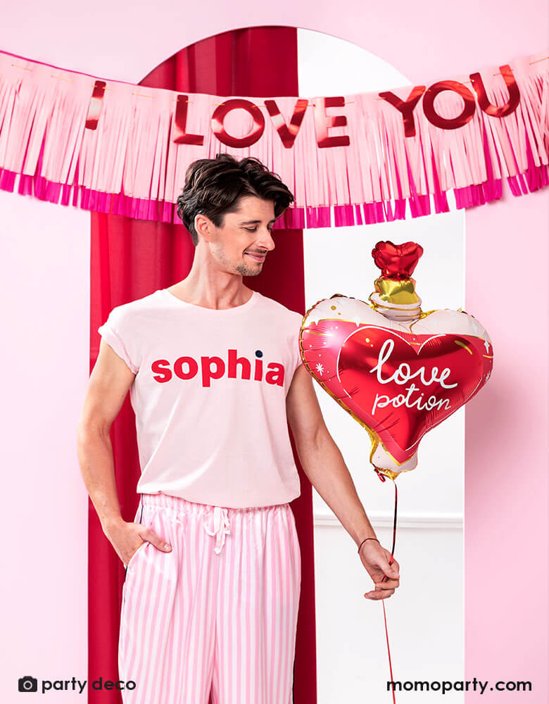 A man dressed in pink pajama holding Momo Party's love potion shaped foil balloon by Party Deco, standing in front of a door decorated with pink tissue-fringe banner with red type message of "I love you" - a perfect way to express your love on Valentine's Day.