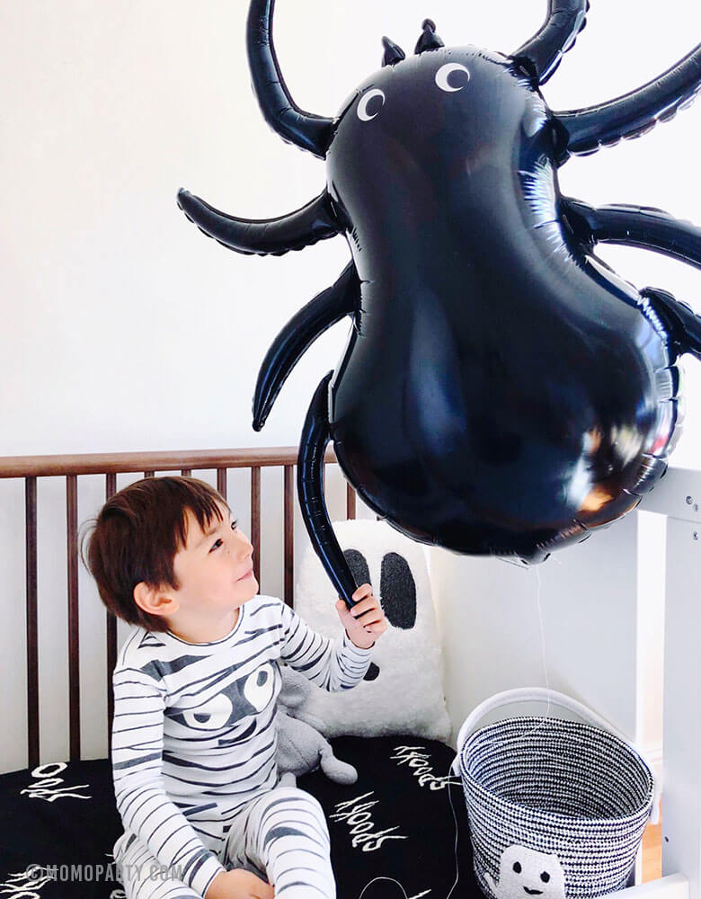 A boy in mummy costume holding a giant Halloween Spider foil Balloon on his bed with Halloween decorations