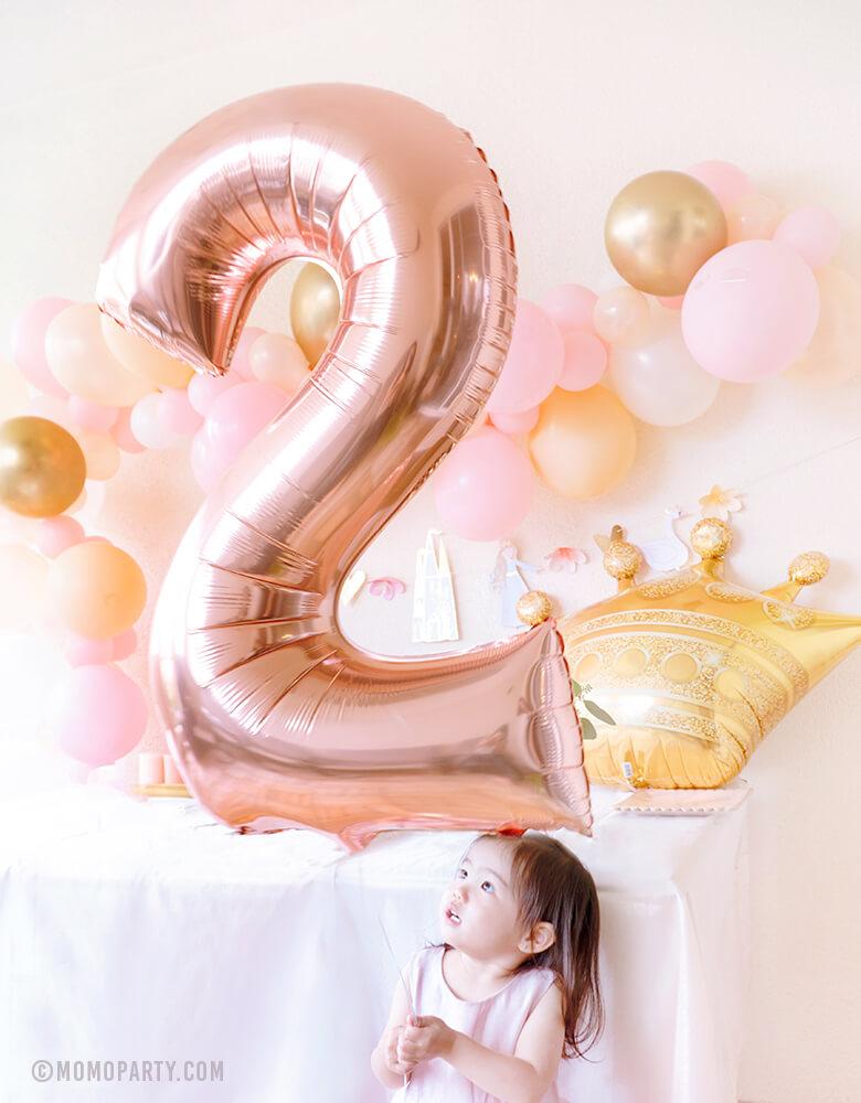A cute girl wearing pink dress and holding a Large Rose Gold Number 2 Foil Mylar Balloon at her 2 years gold and pink princess birthday party