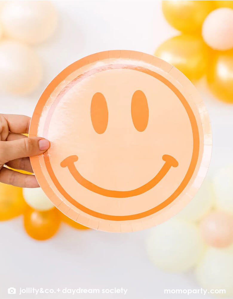 A hand holding Momo Party's 8" peach love and peace smily face dessert plate by Jollity & Co. in front of a wall decorated with daisy balloons. Momo Party's 8" love and peace smily face dessert plate by Jollity & Co. Featuring a groovy, retro design and bursting with boho vibes, these plates will bring good vibes to any girl's birthday.