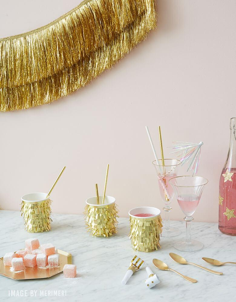 Meri-Meri-Gold-Fringe-Cups with Gold Tinsel Fringe Garland on a pale pink wall for a holiday party