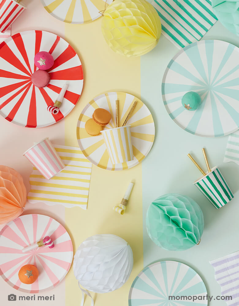 A close up of colorful fun table top filled with Meri Meri Mixed Stripe Dinner Plates and Mixed Stripe Side Plates, Mixed Stripe Napkins, Mixed Stripe paper cups, colorful macarons and honeycomb decoration in the colorful paper tablecloths in rainbow order