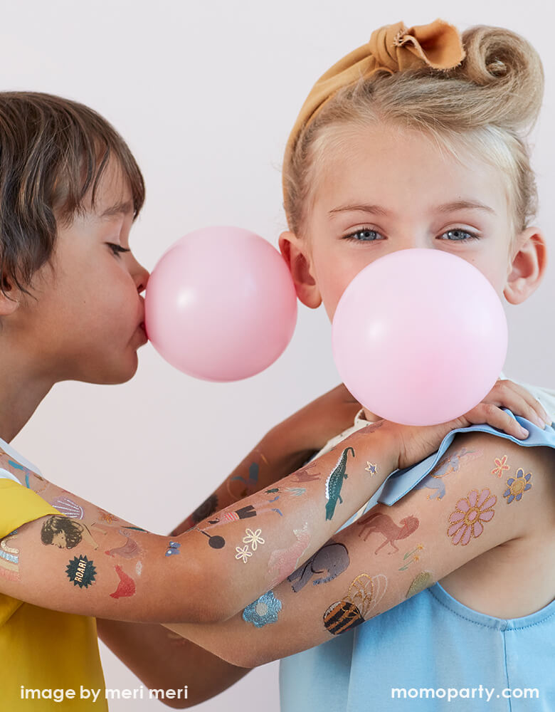 Boy and girl blowing bubble gums and holding each other's shoulder with full of fun bright color and foil detailed temporary Tattoos on their arms