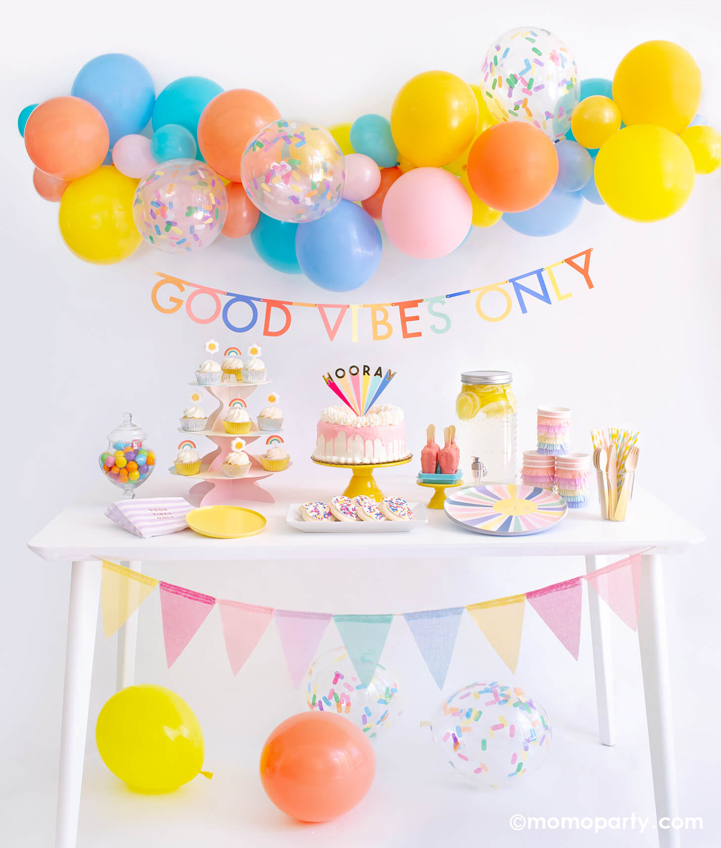 Momo party Good Vibes Only Party Box Kit. Included Meri Meri Rainbow Sun Dinner Plates, Oh Happy Day Yellow Side Plates, Meri Meri Rainbow Sun Fringe Cups, Party Deco Good Vibes Napkins, Yellow Wooden Cutlery, Mixed Pastel Striped Straws, cupcake with rainbow and daisy candles. Meri Meri Hooray cake topper on a dripping cake, colorful gumballs, Lemonade on the table. Meri Meri Multicolor Bunting Banner spelled as "good vibes only" text , Good Vibes 6-foot- Balloon Garland Cloud as decoration.
