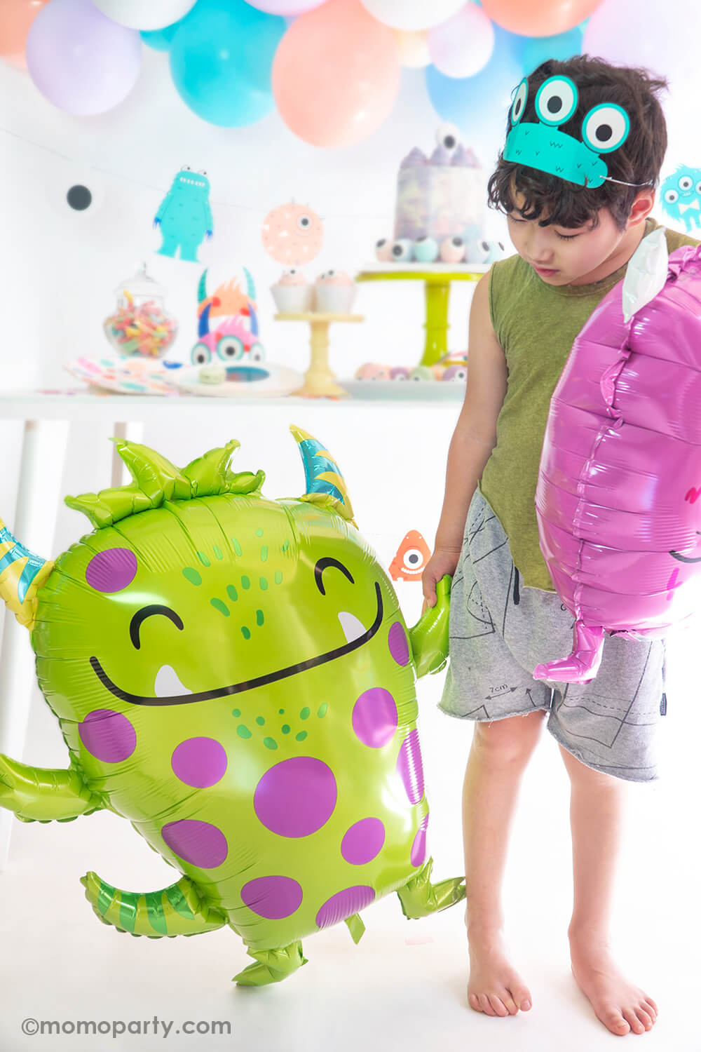 Boy wearing a monster hat, and holding Anagram Balloons - Green Happy Monster Foil Balloon walking in front of the little monster themed birthday party. Also perfect for not so scary halloween party 