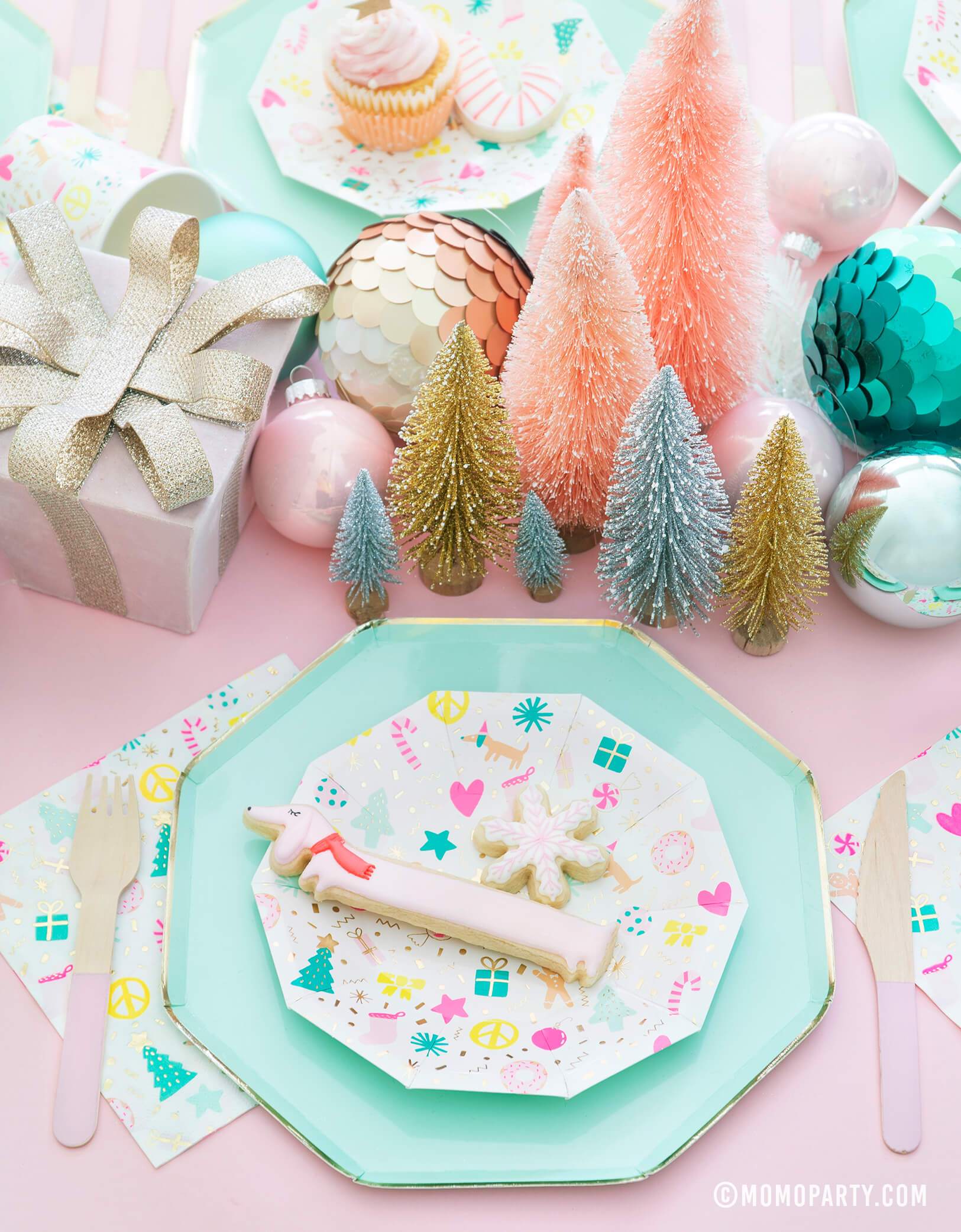 Merry and Bright Pastel Christmas table set up with cookies on top of Meri Meri Mint Large Dinner Plates and Daydream Society Merry and Bright Holiday Christmas Party Plates,Napkins and Cups, Pink wooden utensils, and gift box, Christmas Ornaments, mini pastel Sisal Trees as center piece for a pastel Christmas party celebration    