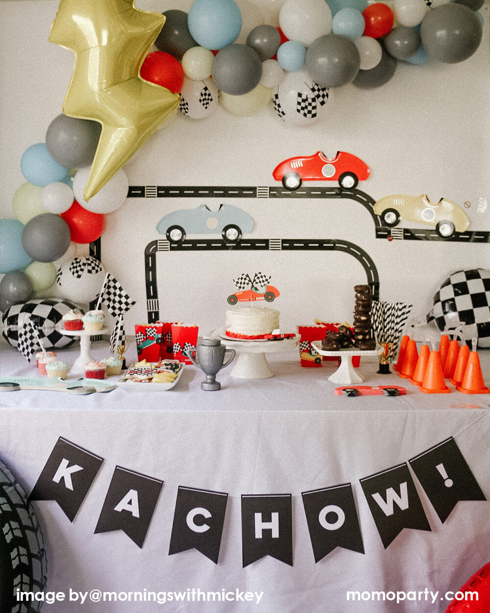 Modern disney race car party, decorated with Road Tape on the wall with Meri Meri Race Car Die-cut Paper Plates, modern pastel themed balloon cloud mix with flag latex balloon and lighting bolt foil balloon, race car topper on a buttercream cake, trophy sipper, mini donuts as tire on a cake stand, red popcorn cups with race car stickers next to the main cake, black letter banner spelled as "kachow!" hanging in front of the table. A modern Car themed Birthday Party at home by Mornings-with-Mickey