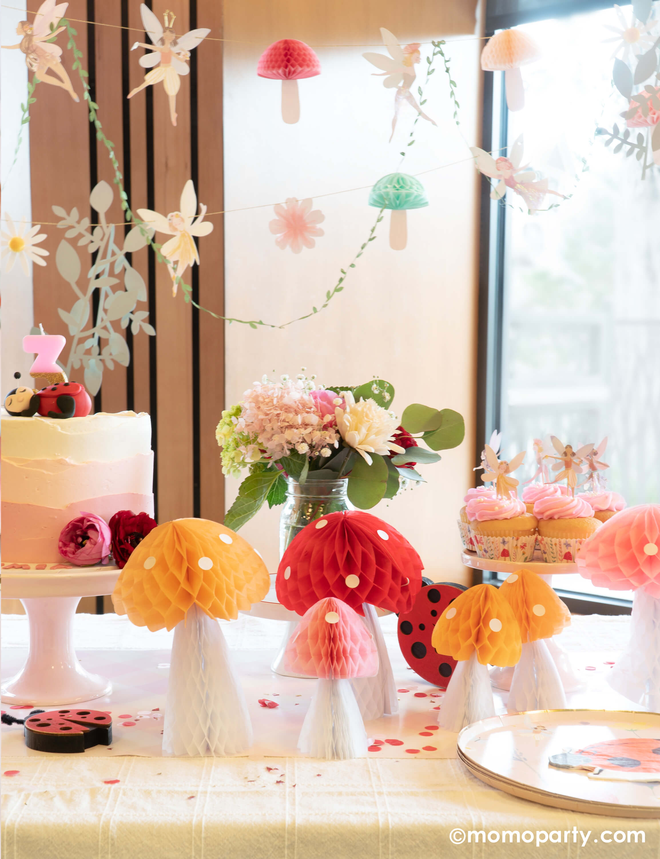 A girl's spring fairy themed birthday party table decoration featuring Momo Party's honeycomb mushroom by Meri Meri and fairy themed partyware including dinner plates, ladybug napkin and fairy cupcake toppers, with a beautiful fairy garland hung above the table, all makes a perfect inspo for a spring party or celebration.