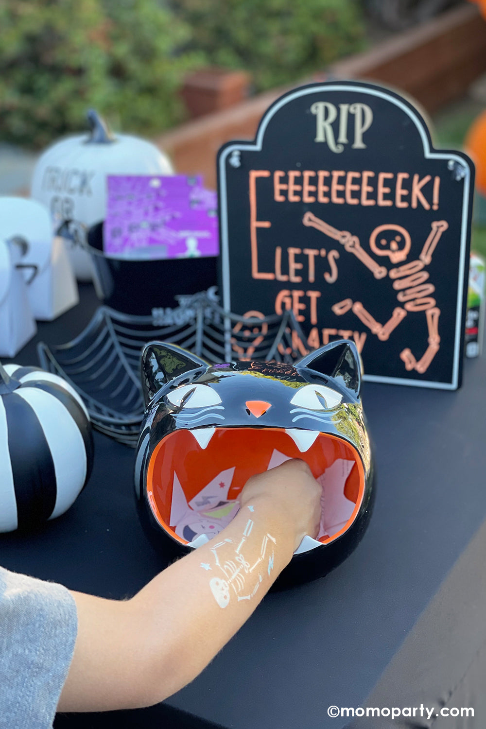 A kid's hand with Meri Meri's Halloween temporary skeleton tattoo on it, trying to grab a treat from a open-month black cat jar in a Halloween birthday party setting. A great kid-friendly Halloween party activity or trick or treat non-sugar treats idea.
