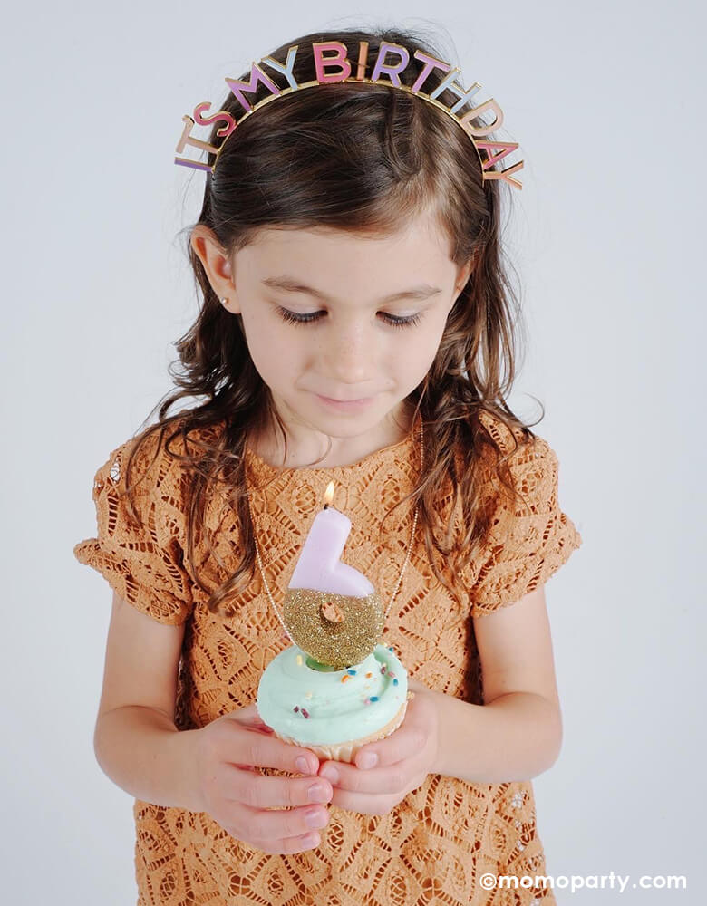 little girl holding a cupcake with number 6 pink gold glitter candle, wearing a Talking Table Rose It's My Birthday Headband, celebrate her six years birthday