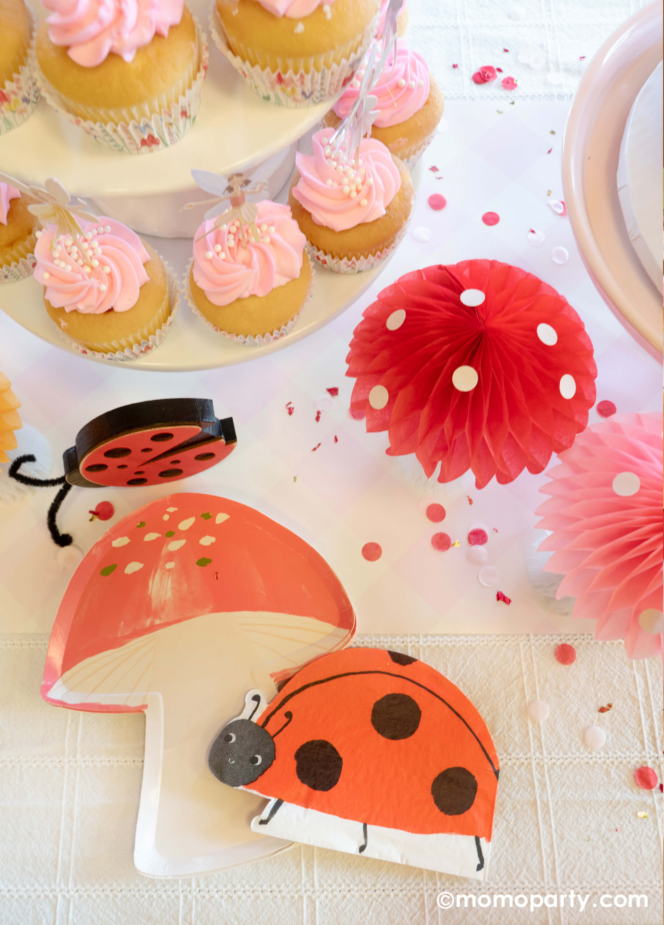 A spring fairy themed birthday party table featuring Momo Party's fairy mushroom shaped plate by Meri Meri, ladybug shaped napkin, mushroom honeycomb decoration next to pink cupcakes topped with Momo Party's fairy cupcake toppers by Meri Meri, all makes a great inspo for a spring inspired party or girl's birthday celebration. 