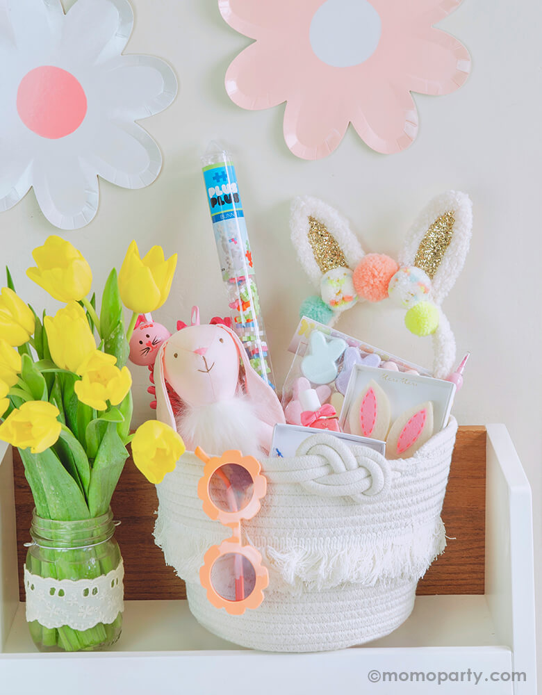 A cute white Woven basket for girl on a bookshelf, there are Pom pom Bunny Ear hairband, Bunny Ear Hair Clips, Pink Bunny Doll, Plus-Plus Easter Bunny Tube, Easter Egg Bunny Bath Bombs, Bunny Jumping Rope, and Snails Washable Nail Polish - Fairytale inside the basket. a peach daisy sunglass hanging on the edge. a yellow tuple flower in a glass jar next to the basket. Daisy flower plates on the wall as wall decoration, so cute and unique for a easter celebration, spring party, girl birthday gift ideas