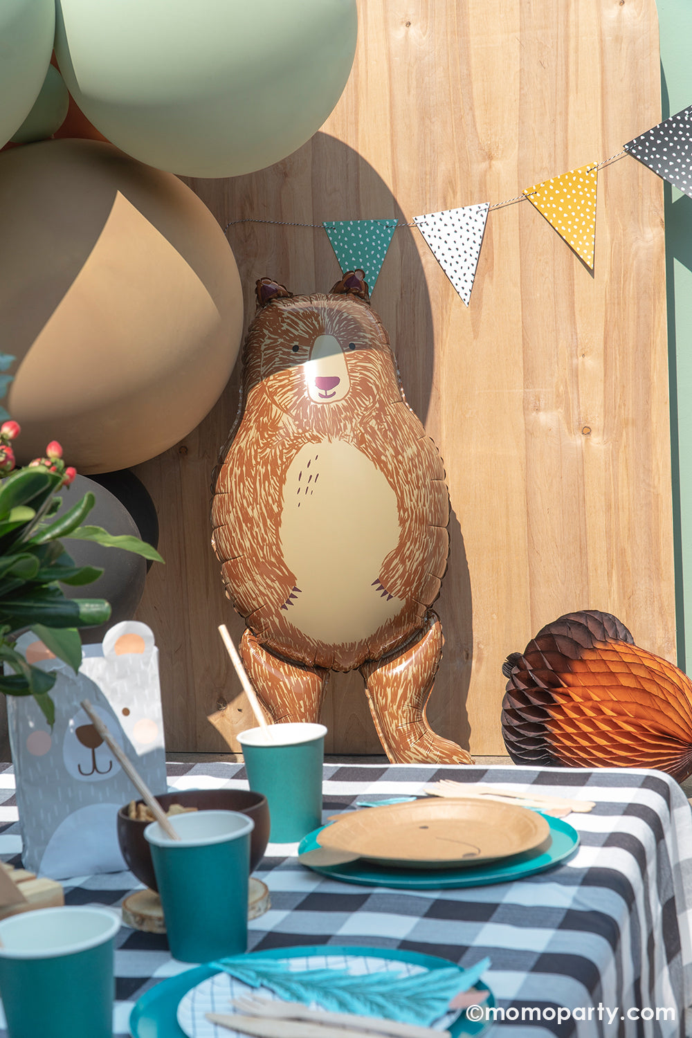 a modern Kid Camping birthday party details with a Honeycomb Acorn decoration next to a Bear foil balloon, in front of a wooden backdrop with Jumbo mocha, green, black latex balloons. In the front there are green cups, bear plates layered oh happy day forest green paper plates, on top of the black and white table cloth.