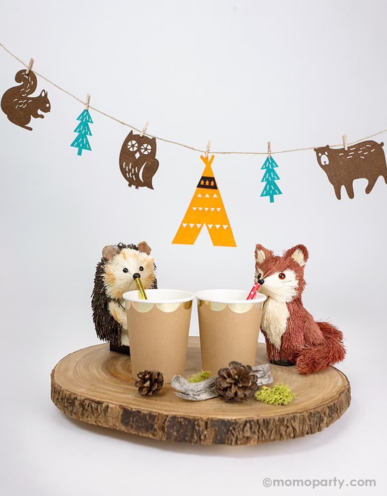 Mini woodland party decorated with a cute Hedgehog and fox figures drinking from a gold scoplated paper cups, with pinecone around them and a woodland felt garland hanging behind them. this woodland felt garland features one teepee, five animals, and three trees. With mini wood clothespins, this set is easy to put together and creates a lovely charm to your woodland celebration