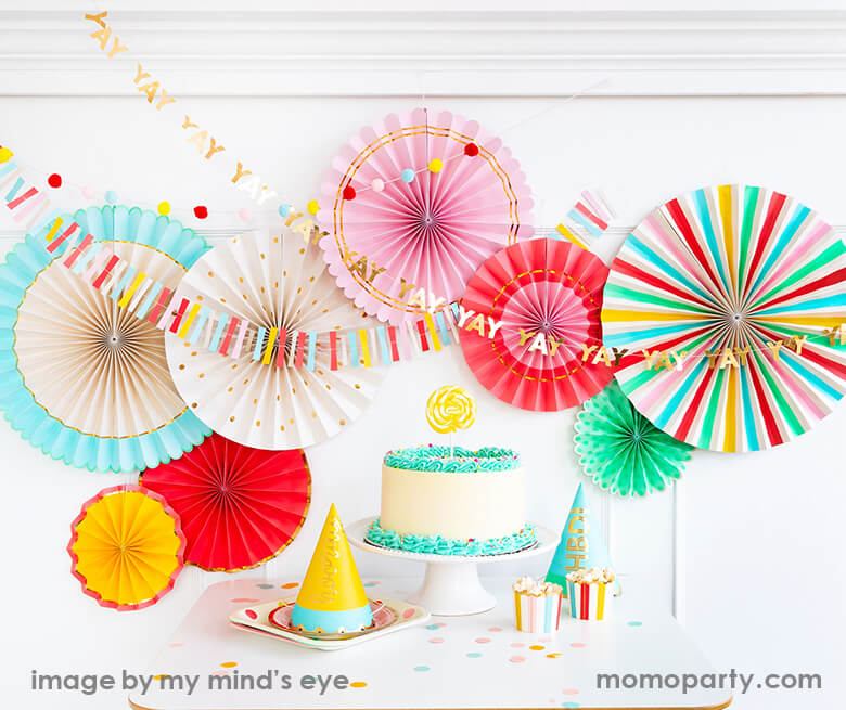 Home party set up with My Minds Eye - Hip Hip Hooray Paper Fans, HIP HIP HOORAY MINI BANNERs decorated on the wall, a buttercream cake with a bright yellow lollipop on it, party hats and popcorns in the colorful stirpe HIP HIP HOORAY FOOD CUPS, bright color confetti around the table for a happy bright party