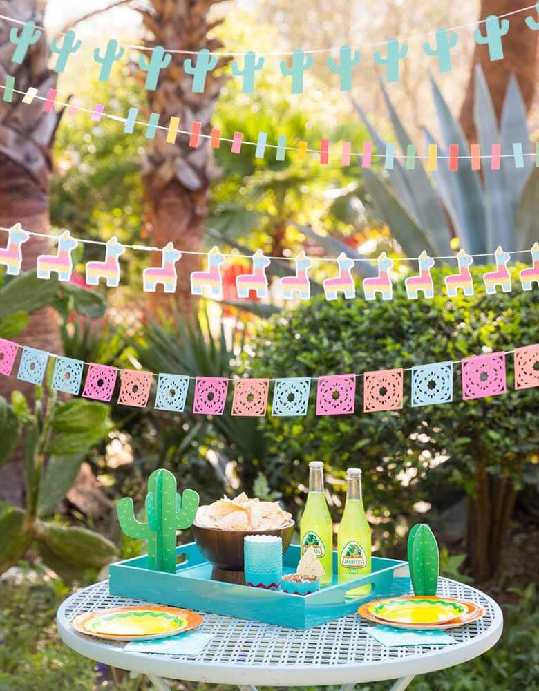 My-Minds-Eye-Fiesta-Mini-Banner-Set with ticker tape banner, llama banner, papel picados banner and mini cactus banner  hung  on top of a party table with cactus decorations for a fiesta themed party