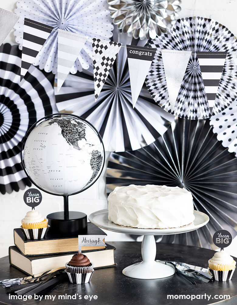 A modern black and white Graduation Party, decorated with My Minds Eye Graduation Party Fan layered with graduation hat Banner on top. a white cake on the cake stand, books, black and white globe as decorations, cupcakes with Graduation cupcakes kit around the table. Celebrate your Class of 2021 grad with this modern graduation collection.