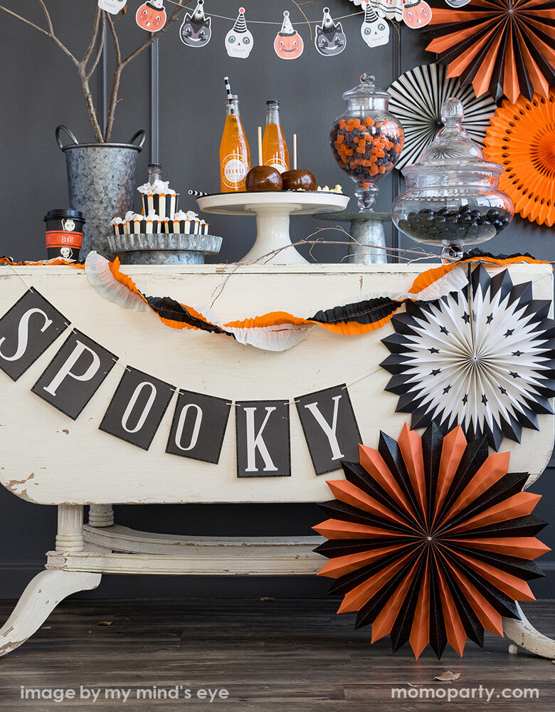 Classic vintage halloween party table look with sweets in My Minds Eye Vintage Halloween Food Cups in the Galvanized metal tray, chocolate apples on top of a cake stand, with candy jars on the side, orange juice in the glass bottle, a Vintage Halloween Paper Fans decorated as backdrop and around the table, a spooky letter banner and halloween vintage crepe paper banner in front of the table,  Such a frightful set up for your haunted house party with these vintage inspired Halloween party collection