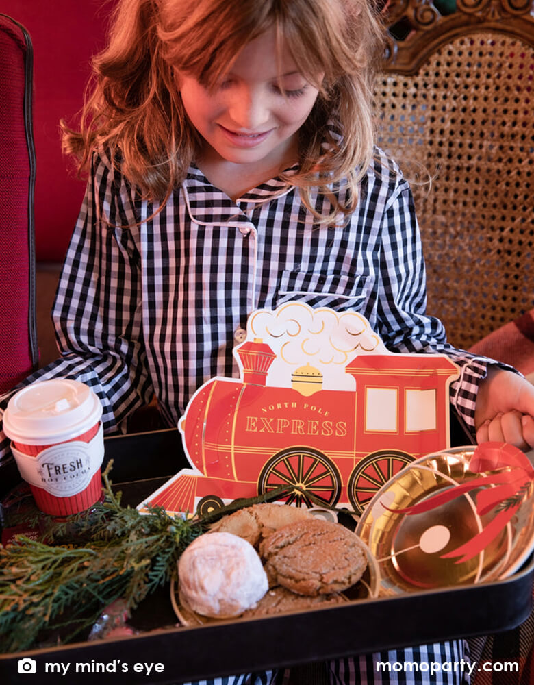 A little girl in her Holiday pajamas holding a vintage tin tray filled with holly branches, holiday treats and cookies and Momo Party's North Pole Express train shaped plates, bell shaped side plates and polar express red 8 oz to-go cups by My Mind's Eye on a vintage Polar Express train cabin red leather seat, ready for a festive Holiday celebration for family and kids.