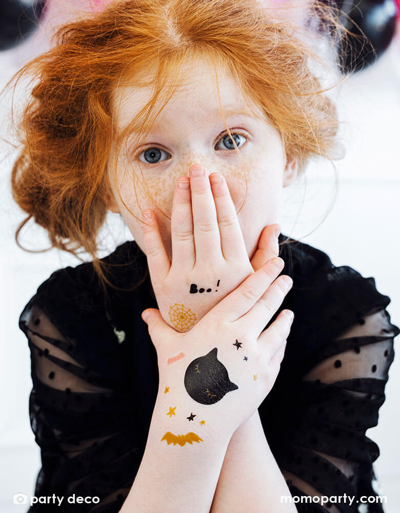 A girl in her black Halloween costume with her arms decorated with Party Deco's Boo Halloween temporary tattoos featuring black cats, spiderweb, bats, and ghost