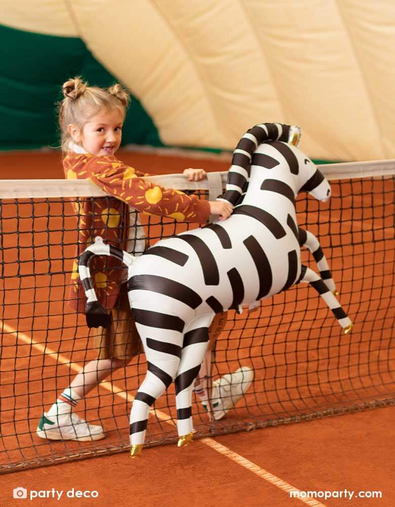 A young preschool aged girl walking along side Party Deco's 45" Zebra Foil Balloon in matte material in an outdoor tennis court