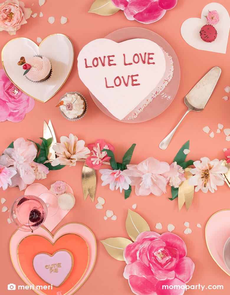 A Valentine's Day themed party table with a heart shaped cake decorated with the message of "love love love" along with Meri Meri's pink tone heart shaped plates and napkins featuring floral garland as the centerpiece of the table, a sweet set up for a Valentine's Day celebration.