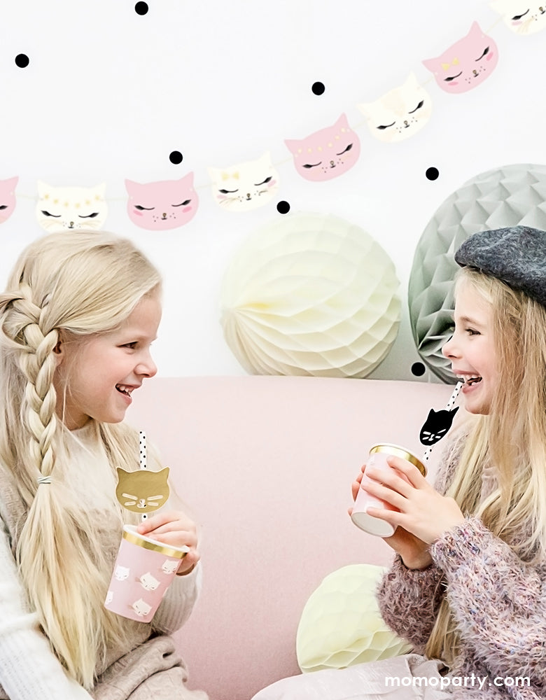 Girls having fun in a kitty cat themed birthday party. They are sitting on the pink sofa, chatting and laughing while drink with Party Deco Kitty cat cups with black dots straws with gold and black cat Paper decorations. A kitty cat garland on the wall, grey and white honeycombs on top of the sofa like a kitten toy. These cat themed party supplies are super cute for a girls birthday, cat lover's birthday, pet themed party
