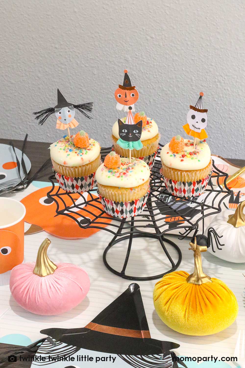 Kid's Halloween party table filled with Halloween themed party supplies including a set of cupcakes decorated with Halloween character toppers including a black cat, a skull, a pumpkin, and a witch on a spider cobweb shaped cupcake stand, creating a spooky vibe for this Halloween season!