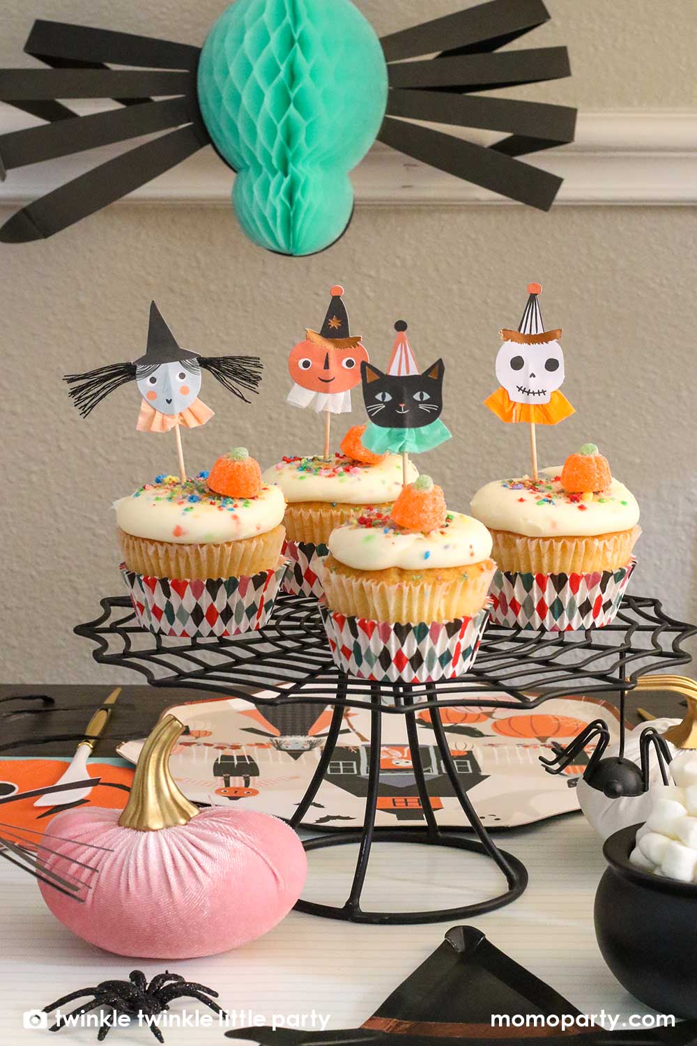 Kid's Halloween party table filled with Halloween themed party supplies including a set of cupcakes decorated with Halloween character toppers including a black cat, a skull, a pumpkin, and a witch. On the table there are Meri Meri's 10inch pumpkin patch dinner plate and happy pumpkin shaped napkins with googly eyes, on the top there was a honeycomb spider decoration hanging from the ceiling, creating a spooky vibe for this Halloween season!