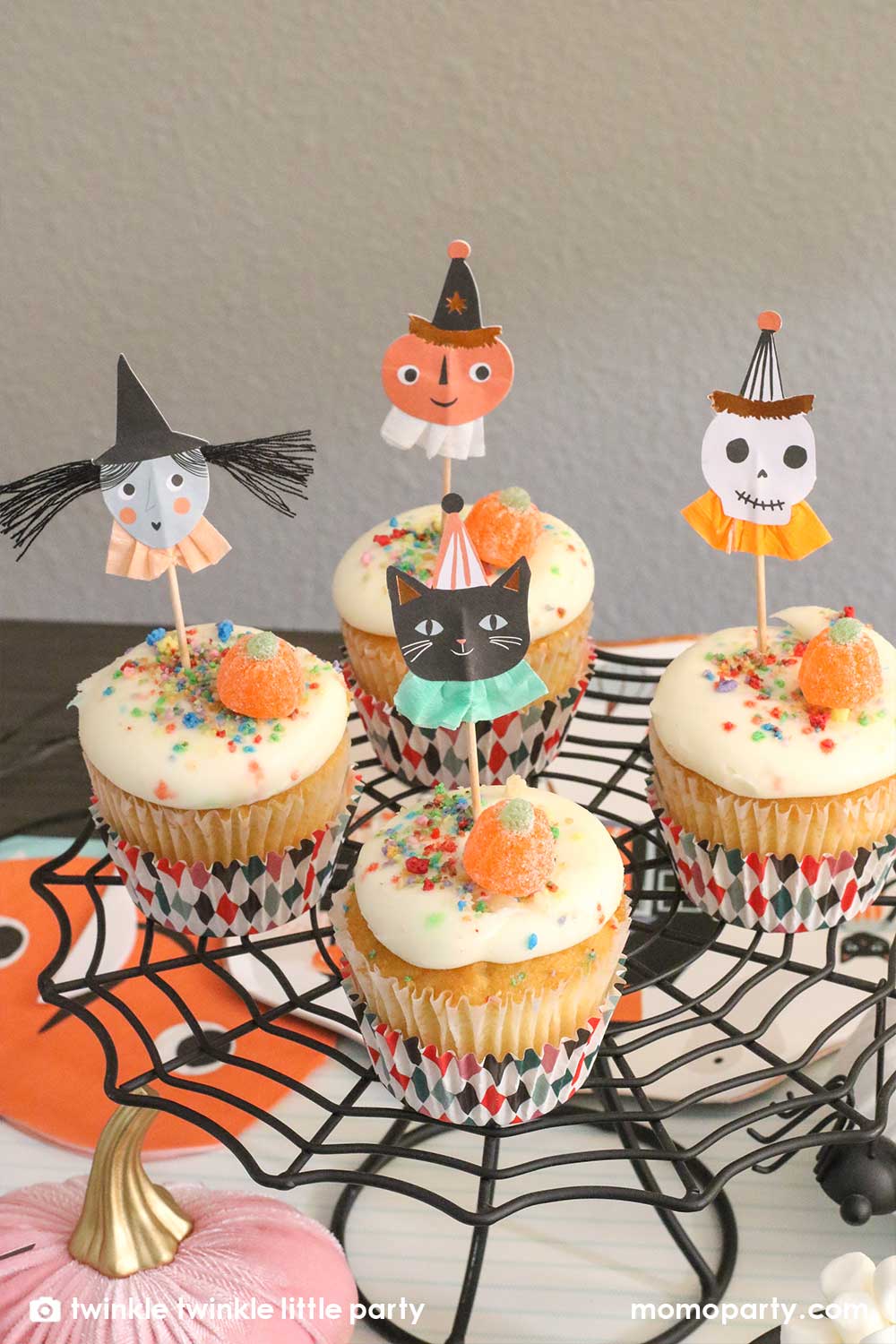 Kid's Halloween party table filled with Halloween themed party supplies including a set of cupcakes decorated with Halloween character toppers including a black cat, a skull, a pumpkin, and a witch on a spider cobweb shaped cupcake stand, creating a spooky vibe for this Halloween season!