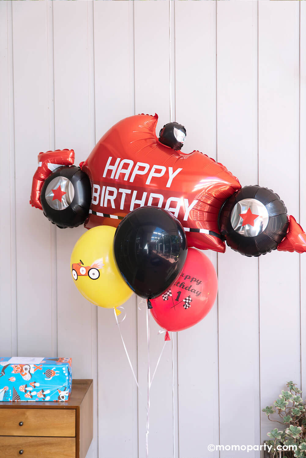 Race Car themed birthday Party corner a wrapped gift next to a Betallic Balloon 43 inch Race Car Birthday Foil Balloon and Vintage Race Car Latex Balloon Mix balloon boutique. Accent your little one's race car birthday party with this awesome car shaped foil mylar balloon with with "happy birthday" text print, and a vintage car print on a yellow latex balloon and a race car flag and "happy birthday" text on a red latex balloon